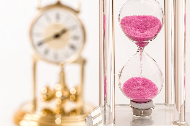 A blured wall golden clock on the left while the focus is on the pink hourglass on the right