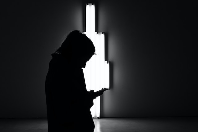 A woman scrolling her phone's silhouette and a light behind in a dark room
